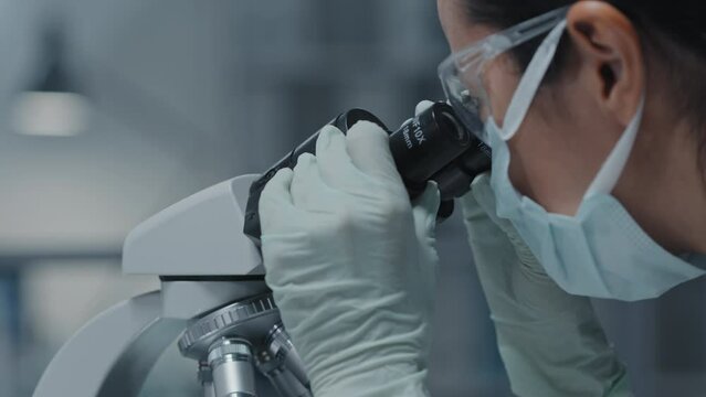 Side view close-up of young female lab assistant in protective goggles, gloves and mask using microscope