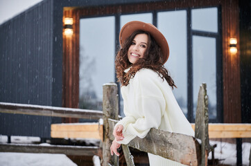 Curly young woman resting near modern barn house in the mountains. Happy female tourist enjoying winter holidays in new cottage.