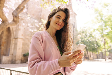 Bottom view of nice young caucasian woman looking at camera uses her phone in sunny weather. Brunette wears singlet and pink cardigan. Concept gadgets.