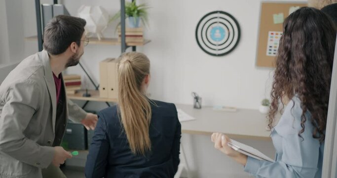 Excited businesswoman is playing darts in workplace relaxing laughing with carefree colleagues. Modern business and youth lifestyle concept.