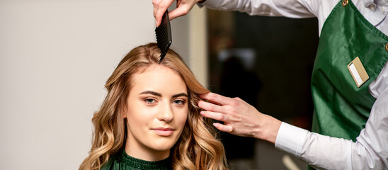 Hairdresser combing long hair of young caucasian woman looking at the camera and smiling in beauty salon