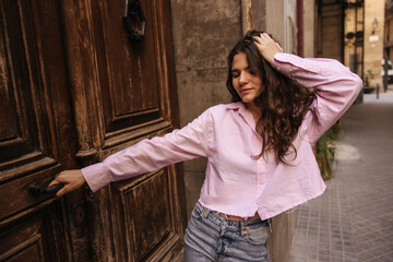 Fototapeta premium Pretty young caucasian brunette woman posing near old door with closed eyes spends time outside. Brunette wears pink shirt and jeans shorts. Concept urban, street style.