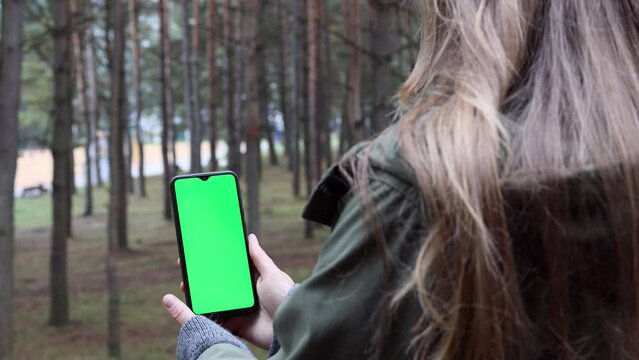 Smartphone with a green screen against the background of forest and trees and bushes.
Woman holding vertical mobile phone with green mock up screen. Concept of using technology in nature.