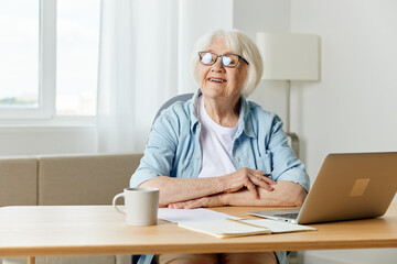 a happy elderly woman with gray hair is sitting at her desk with a laptop and smiling broadly looks away with her hands folded on the table. The concept of working from home