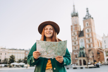 Attractive young female tourist is exploring new city. Redhead girl in hat holding a paper map on Market Square in Krakow. Traveling Europe in autumn. St. Marys Basilica