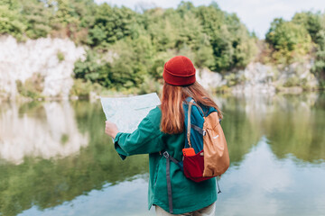 Back view of stylish hipster woman holding paper map, wearing backpack and red hat looking at...
