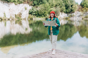 Stylish hipster woman holding paper map, wearing backpack and red hat looking at mountain view while relaxing in nature. Travel and wanderlust concept. Amazing chill moment. Full body photo
