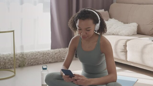 Slowmo of Biracial girl in tight activewear enjoying music in headphones while sitting on mat at home, resting between exercises