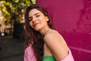 Peaceful young caucasian woman looking at camera smiling, stands outdoor during spring day. Brown hair girl wears casual clothes. Concept solo vacation.