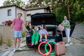 Happy father holding power supply cable and charging their electric car, rest of family putting suitcases in car trunk, preparating for holidays.