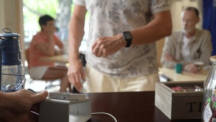An elder man pay the bill with smart watch wirelessly on POS terminal using NFC contactless technology in a cafe. Purchase by smartwatch on electronic payment machine. E-money at cashless wallet. - Powered by Adobe