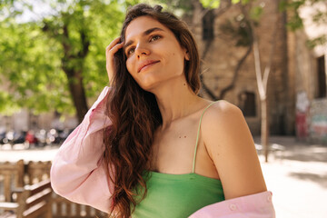 Cool young caucasian girl stands at european street alone, collects her hair with hand. Woman with brown wavy hair wears green singlet and pink shirt. Concept solo vacation.