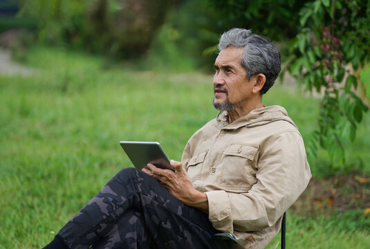 A Senior Man Resting On Camping Chair, A Male Older Adult Thinking While Using Tablet Computer In The Garden,concept Elderly People Lifestyle, Hobby, Small Business Owner In Farm, Casual Business