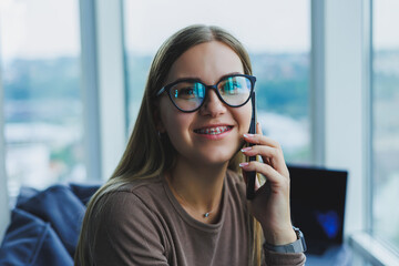 Young smiling woman wearing glasses in casual clothes sitting in a chair by the window and talking on the phone during a video call.