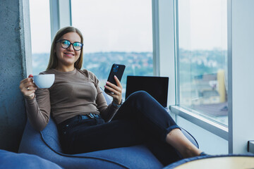 Blonde young smiling woman in glasses in casual clothes sitting in a chair by the window with a phone and drinking coffee