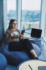 Blonde young smiling woman in glasses in casual clothes sitting in a chair by the window with a phone and drinking coffee