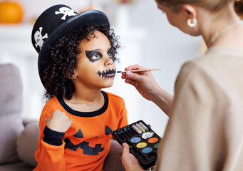 Festive makeup for Halloween. Woman doing pumpkin make-up for ethnic curly boy in costume while...