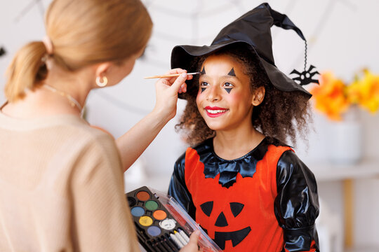 Festive makeup for Halloween. Woman doing witch make-up for ethnic curly girl in costume while preparing holiday