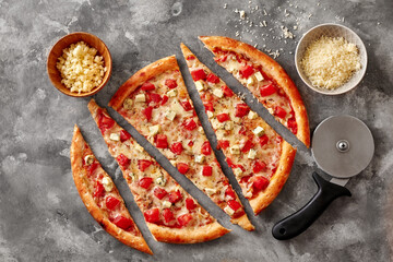 Sliced pizza with tomatoes and blue cheese on stone surface with cutter and bowls of grated...