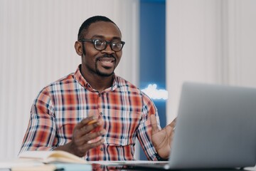 Smiling puzzled african american man reading email with news, working at laptop, spreading hands