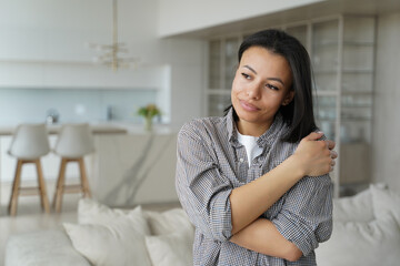 Calm happy female embraces herself feeling self love, self-pride, dreaming, standing at cozy home