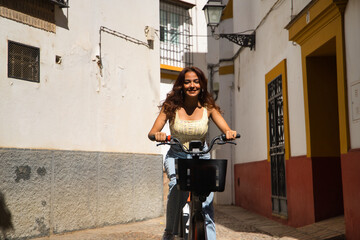 Young beautiful woman with long brown hair and cinnamon skin riding a rented electric bicycle, very happy and content on a trip in a Mediterranean city. Beauty concept, travel, urban mobility.