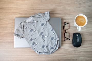 Baby onesie, coffee, glasses laptop on wooden table indoors symbolising maternity or parental leave