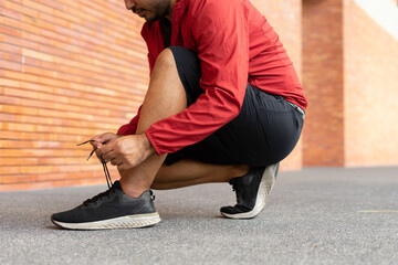 Closeup of sportsman wearing a red jacket tying sneakers outdoors brick wall on city street. Athletic male in hoodie sweatshirt red stopping lacing shoes in urban city park.