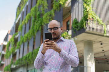 Bearded businessman smiling wearing glasses showing modern smartphone device typing text message in social media stanting front modern building . Happy male spending free time in break of working.