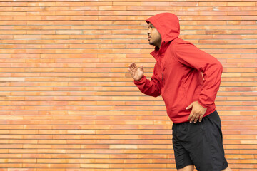 Sportsman wearing a red jacket running on the background of a brick wall stadium. Fitness man runner on urban street. Male jogging and exercise outdoor. Asian athlete man training run on city street.