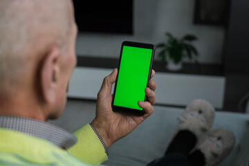 Senior man seated on sofa at home holding vertically mobile device with blank green screen for mockup