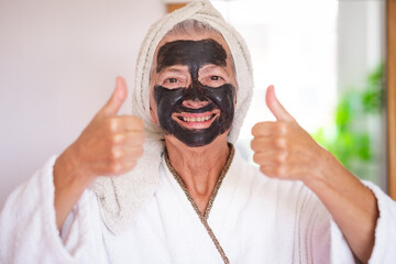 Carefree senior beautiful woman with a detox facial charcoal mask homemade smiling looking at camera with thumb up - take care of the skin concept