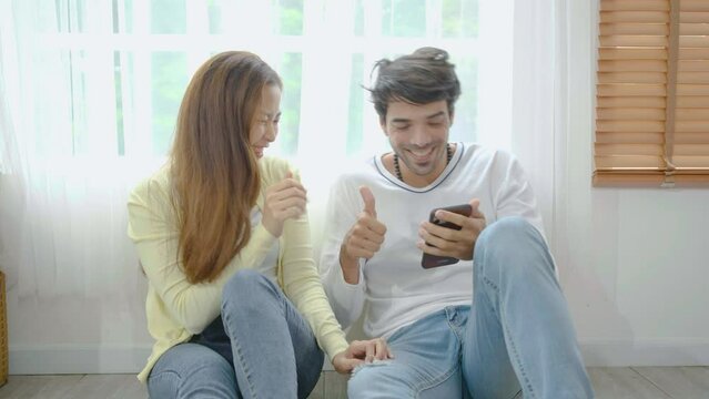 Relaxed young interracial couple or friends using a generic mobile phone together sitting in the living room at home, Happy young man and woman with smartphone, glad to work successfully