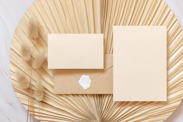 Blank cards and envelope on dry beige palm leaf near hare's tail grass top view,  boho wedding mockup