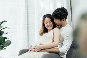 Beautiful pregnant asian couple are sitting on a couch in the living room. The husband is embracing his wife from the back with smiles on their faces and rubbing his wife's tummy.