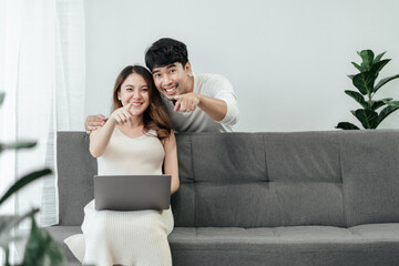Obraz premium Beautiful pregnant asian couple are sitting on a couch in the living room, looking and pointing at camera with smiles on their faces. Happiness Asian couple use computer laptop planing for the future.