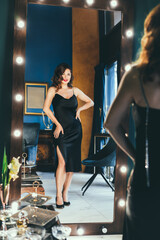 Naklejka na ściany i meble Sexy young woman in elegant cocktail silk black dress dancing and posing in mirror reflection in luxury interior. Fashionable and self-confident lady. Celebrity, superstar lifestyle. Vertical card