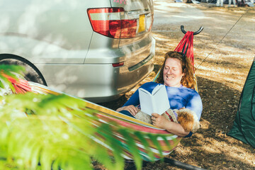 Adult woman relaxing and reading book while lying in hammock with cockapoo puppy near motorhome on camping trip. Female living on camper car with animals and travel the world. Caravan car Vacation.