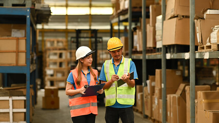 Warehouse workers wearing safety hardhat discussing work while walking in aisle between rows of tall shelves full of packed boxes