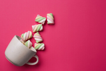 Overturned cup with delicious colorful marshmallows on crimson background, flat lay. Space for text