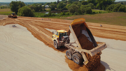 Industrial dumper truck working on a highway construction site, unloading soil. Bulldozer leveling...