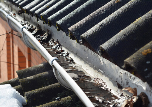 Roof gutter cleaning. A clogged rain gutter of an asbestos roof with non flowing water from melted snow because of unclean gutters with dirt and fallen leaves. Rain gutter before cleaning.