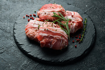 Raw cutlets wrapped in bacon served on a black plate with rosemary and thyme. On a stone background. Top view.