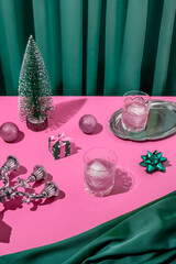 Pacific pink retro futuristic Christmas table arrangement with a tree, gift, bow, pink ice cold cocktails in crystal glass with emerald green curtain background. New Year's eve dinner party aesthetic.