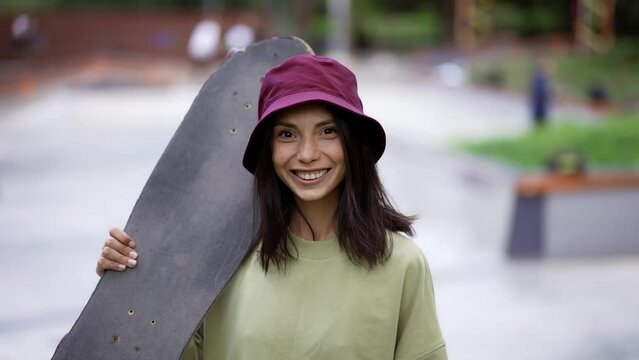 Portrait of a young brunette girl on the background of the park. The girl has a skateboard in her hands, and a red stylish hat is on her head. Teenage hobbies, Leisure