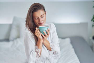 Brunette woman on bad in pajama with mug with coffee or tea