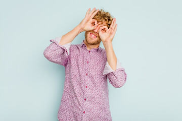 Young caucasian man isolated on blue background showing okay sign over eyes