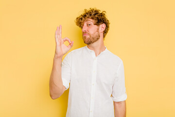 Young caucasian man isolated on yellow background winks an eye and holds an okay gesture with hand.