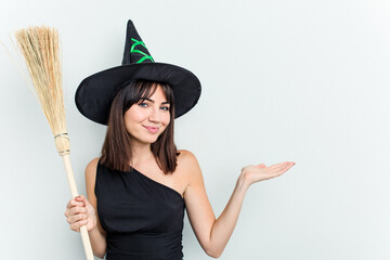 Young caucasian woman dressed as a witch holding a broom isolated on blue background showing a copy space on a palm and holding another hand on waist.