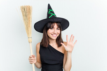 Young caucasian woman dressed as a witch holding a broom isolated on blue background smiling cheerful showing number five with fingers.